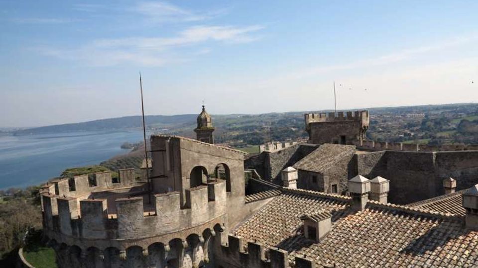 Veduta dall’alto di un castello in pietra con mura merlate, torri e tetti in coppi, con cortile interno e arcate. Sullo sfondo si estendono un lago e colline rurali sotto un cielo sereno.