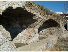 Resti di strutture in muratura con grandi archi in mattoni incassati in un pendio roccioso, affacciati su un’area pavimentata con vasche rettangolari. Sullo sfondo si vede cielo sereno e alcune costruzioni moderne in cima alla scarpata.