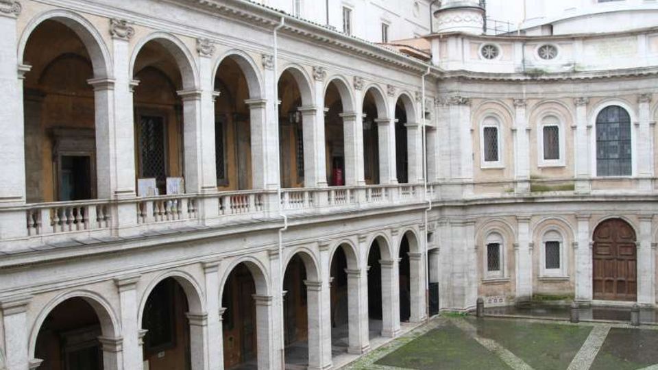 Cortile interno in pietra con portici su due livelli, scanditi da archi e colonne, e balaustra al piano superiore. Sullo sfondo si vede una facciata curva con finestre ad arco e un grande portone, affacciata su un pavimento a motivi geometrici.