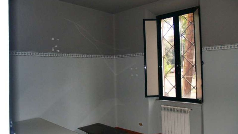 Interno di una piccola stanza d’ufficio con pareti grigie e una fascia decorativa, illuminazione a neon a soffitto e pavimento grigio. Sulla destra c’è una finestra a battenti con inferriata e radiatore sotto, mentre nell’angolo si vedono una scrivania e un tavolino basso.
