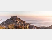 Borgo collinare con case in pietra addossate tra loro e un grande castello fortificato in cima. In lontananza si vedono colline e una valle coperta da nebbia sotto un cielo chiaro.