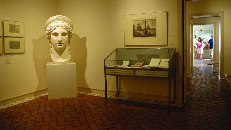 Sala museale con pavimento in mattoni disposti a spina di pesce, una grande testa scultorea su piedistallo e una teca in vetro con documenti; alle pareti sono appesi quadri incorniciati e si intravede un corridoio verso un’altra stanza.