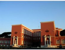 Edificio istituzionale in stile razionalista con facciata rosso mattone, due corpi laterali simmetrici e finestre verticali su più piani. All’ingresso ci sono nicchie con statue e un’ampia area frontale aperta sotto un cielo sereno, con alberi sullo sfondo.