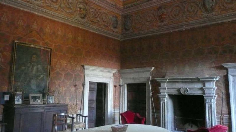 Interno di una sala storica con pareti affrescate e soffitto a cassettoni decorato, dotata di grande camino in pietra con cornice scolpita e porte con stipiti bianchi. Al centro c’è un tavolo rotondo coperto da tovaglia, con sedie e poltrone rosse, e un grande dipinto appeso sopra una boiserie.