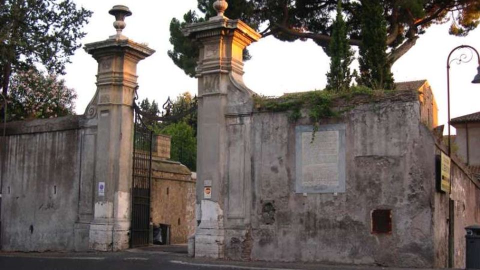 Ingresso con cancellata in ferro tra due alti pilastri in pietra sormontati da vasi ornamentali, inserito in un lungo muro intonacato e invecchiato. Sullo sfondo si vedono alberi alti e vegetazione sopra il muro, con strada asfaltata in primo piano.