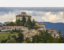 Veduta panoramica di un borgo collinare con edifici in pietra e tetti in coppi, dominato da un grande palazzo o castello su un’altura circondata da alberi. Sullo sfondo si vedono colline boscose e un cielo nuvoloso.