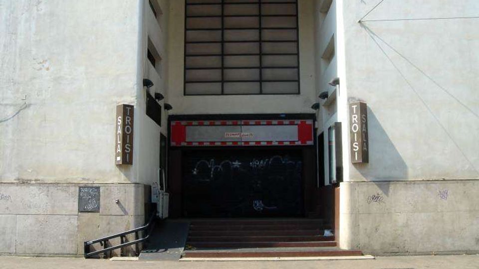 Ingresso incassato in un edificio chiaro con grande finestra rettangolare a griglia sopra e portone scuro chiuso, raggiunto da una breve scalinata e una rampa laterale. Ai lati dell’entrata sono presenti insegne verticali e pareti in muratura con segni di usura.