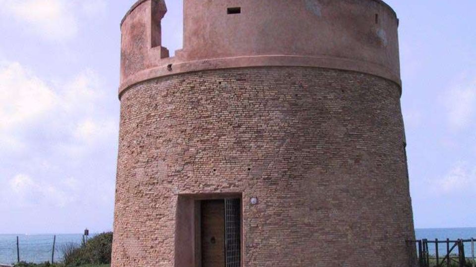 Torre costiera cilindrica in mattoni, con coronamento intonacato e piccola apertura in alto, vista frontalmente. All’ingresso si nota una porta con grata; la struttura sorge su terreno spoglio vicino al mare, con recinzione e cielo aperto sullo sfondo.