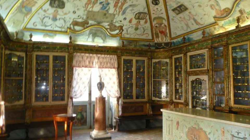 Interno di una sala storica con soffitto a volta affrescato e pareti rivestite da grandi vetrine lignee ornate, piene di piccoli oggetti esposti. Al centro si vede una finestra con tende leggere e un piedistallo, mentre in primo piano c’è un bancone decorato e il pavimento in piastrelle.