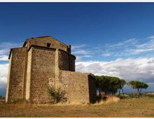 Chiesa in pietra di aspetto romanico con abside semicircolare e volume massiccio, situata in un’area rurale con terreno erboso secco. Sullo sfondo si vedono file di pini e un ampio cielo azzurro con nuvole.