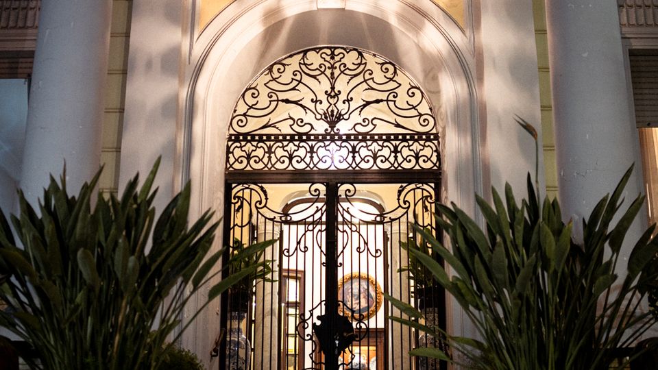 Ingresso di un edificio classico con due alte colonne e un portale ad arco, chiuso da un cancello in ferro battuto decorato e illuminato da una lanterna sospesa. Ai lati della scalinata ci sono grandi vasi con piante, con l’interno visibile in controluce oltre il cancello.