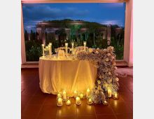 Tavolo rotondo con tovaglia bianca allestito con candele in cilindri di vetro e decorazioni floreali, con un’ampia composizione di fiori bianchi che scende sul lato fino al pavimento. Sullo sfondo, una grande vetrata affaccia su un giardino con alberi e colonne illuminate al crepuscolo.