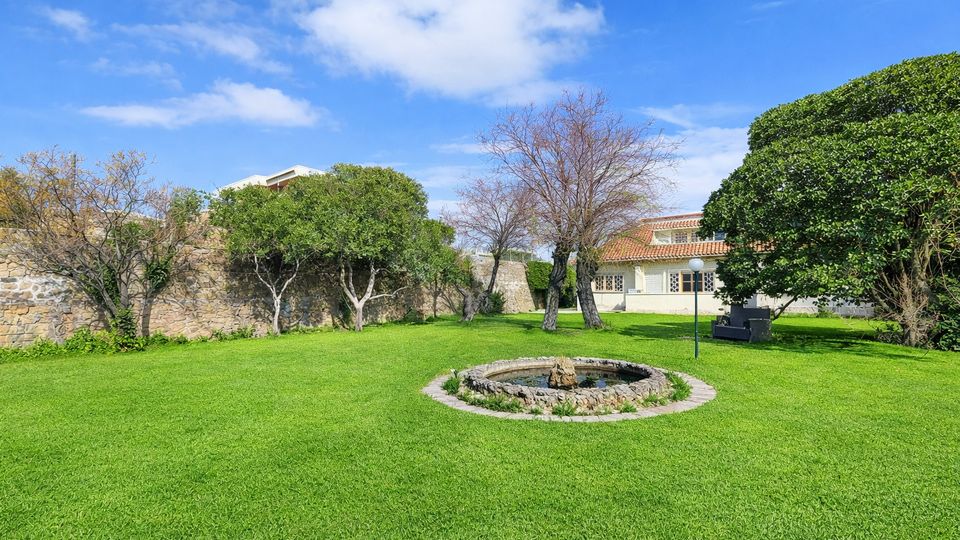 Ampio giardino con prato verde e una piccola fontana circolare in pietra al centro, circondato da alberi e cespugli. Sullo sfondo si vedono un muro in pietra e un edificio con tetto in tegole rosse sotto un cielo azzurro con nuvole sparse.