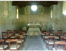 Interno di una piccola cappella in pietra con soffitto a travi in legno, navata centrale con sedie in legno e altare rivestito da tovaglia bianca. Sul fondo si vede un grande oculo circolare sopra l’altare, con affreschi sbiaditi e strette finestre laterali.