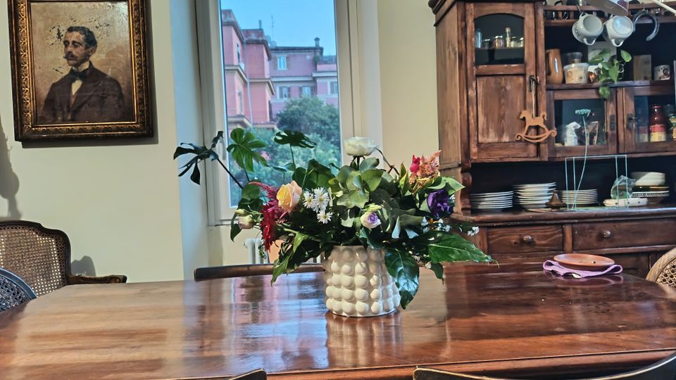 Sala da pranzo domestica con grande tavolo in legno lucido e vaso di fiori al centro, affiancata da un’ampia finestra che dà su palazzi e alberi. Sullo sfondo si vede una credenza in legno con piatti e tazze a vista e, sulla parete, un quadro incorniciato.