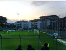 Campo da calcio di uno stadio di quartiere con tribune laterali recintate e illuminazione a torre; sullo sfondo si vedono palazzine residenziali e un cielo al tramonto. In primo piano sono visibili la rete metallica di separazione e un’area vicino alla porta con bandierina d’angolo.