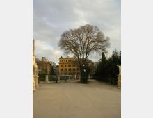 Viale ampio e sterrato di un parco urbano, fiancheggiato da statue in pietra su piedistalli e lampioni, con un grande albero spoglio al centro. Sullo sfondo si vedono palazzi in stile storico e aree alberate ai lati sotto un cielo nuvoloso.