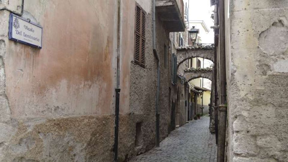 Vicolo stretto con pavimentazione in ciottoli tra edifici in pietra e intonaco deteriorato, con finestre dotate di persiane e tubi di scolo a vista. In fondo si vedono un arco di collegamento tra gli edifici e un lampione, con una targa stradale sul muro a sinistra.