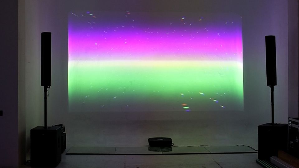 Interno di uno spazio espositivo industriale con pavimento in cemento e soffitto a travi, con un proiettore sospeso. Su una parete bianca è proiettata un’immagine rettangolare a bande colorate viola e verde, affiancata da due altoparlanti su supporti.