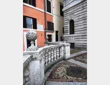 Vicolo urbano tra edifici storici con facciate color ocra e arancione e un palazzo in pietra bugnata con finestra ad arco. In primo piano un parapetto in travertino con balaustre e sfere decorative, accanto a una pavimentazione in pietra e mattoni con motivo circolare.