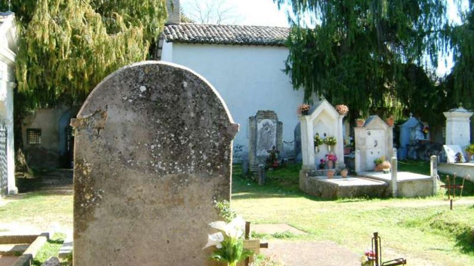 Piccolo cimitero all’aperto con lapidi e tombe in pietra, alcune con croci e fiori, su un prato. Sullo sfondo si vede un edificio bianco con tetto in tegole, circondato da alberi alti.