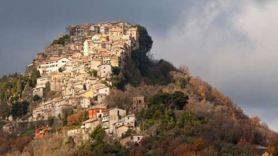 Borgo collinare con case in pietra e intonaco addossate tra loro, arroccato sulla sommità di un rilievo boscoso. Sullo sfondo un cielo nuvoloso, con vegetazione autunnale che ricopre i versanti.