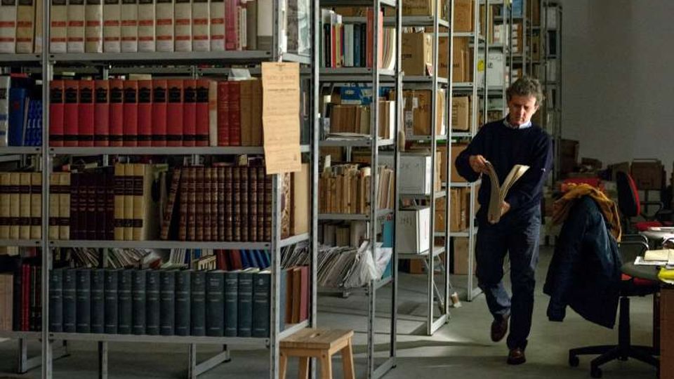Interno di un archivio o biblioteca con alte scaffalature metalliche piene di volumi rilegati, fascicoli e scatole etichettate. Sul lato destro si vede una postazione di lavoro con sedia girevole e scrivania, in un ambiente illuminato da luce naturale.