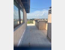 Terrazza sul tetto con pavimento piastrellato e parapetti in muratura, affiancata da una parete con grandi finestre e sporgenza del tetto. In fondo si vede una fioriera con vegetazione e un panorama urbano con tetti e cielo azzurro.