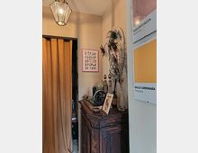 Angolo di un interno con pareti beige, una lanterna a soffitto e una tenda pesante che copre un’apertura. Accanto, un mobile in legno scuro con oggetti decorativi e un vaso con piume di pavone, mentre sulla parete sono appesi riquadri e pannelli con campioni di colore.
