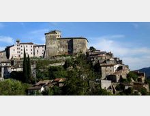 Veduta panoramica di un borgo su collina con un grande castello in pietra e torre quadrata in cima, circondato da edifici storici compatti con tetti in coppi. In primo piano si vedono alberi e vegetazione, con il profilo delle colline sullo sfondo sotto un cielo sereno.
