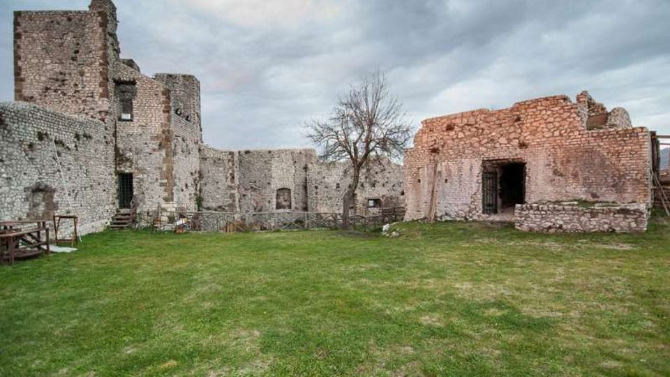 Cortile erboso all’interno di un complesso di rovine in pietra, con mura alte e aperture ad arco tipiche di una struttura fortificata o castello. Sullo sfondo si vedono un albero spoglio, recinzioni basse e un cielo nuvoloso.