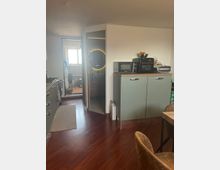Interno di una cucina moderna con pavimento in legno scuro, mobili verde salvia e piano in legno con microonde e piccoli elettrodomestici. A sinistra c’è una fila di basi cucina e, in fondo, una porta conduce a un bagno con specchio rotondo illuminato e a una zona luminosa con finestra.