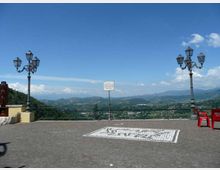 Piazzale panoramico pavimentato in pietra con ringhiera affacciata su una valle e colline lontane. Due lampioni in ferro battuto e un cartello centrale incorniciano la vista sotto un cielo azzurro.