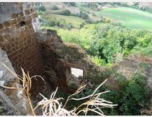 Rovine di una struttura in muratura di mattoni e pietra, con pareti parzialmente crollate e aperture rettangolari. Sullo sfondo si vede una vallata con boschi e campi coltivati.