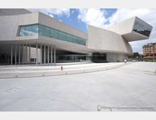 Edificio moderno in cemento chiaro con volumi sporgenti e grandi vetrate orizzontali, sostenuto da una fila di pilastri sottili. Davanti si estende un’ampia piazza pavimentata con gradinate basse e aiuole, sotto un cielo parzialmente nuvoloso.