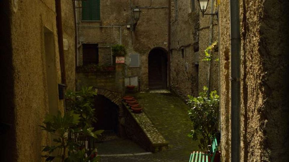 Vicolo stretto in un borgo storico con pavimentazione in ciottoli, fiancheggiato da edifici in pietra con persiane e lampioni a muro. In fondo si vede un piccolo ponte ad arco e una rampa con vasi di piante; sul lato destro c’è una panchina verde vicino alla parete.