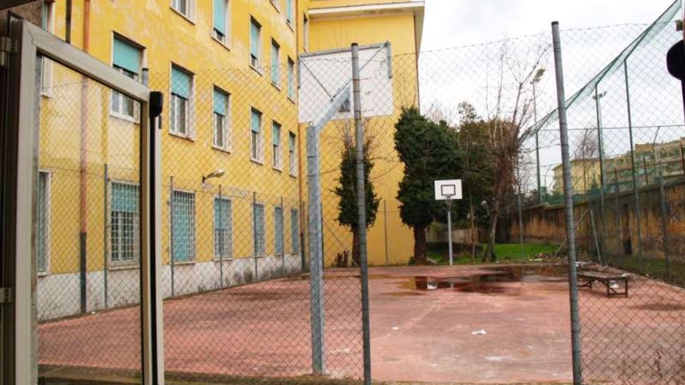 Cortile esterno recintato con campo da basket in cemento rosso, canestro e panchina, affiancato a un grande edificio giallo a più piani con finestre e grate. Sullo sfondo si vedono alberi e una piccola area verde oltre la rete metallica.
