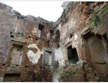 Ruderi di una struttura antica in pietra e tufo, con pareti parzialmente intonacate e aperture rettangolari e ad arco scavate nei muri. L’ambiente è a cielo aperto, con segni di erosione e vegetazione che cresce tra le pietre.