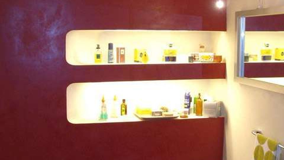 Bagno moderno con parete bordeaux e nicchie orizzontali illuminate usate come mensole per prodotti. A destra si vedono uno specchio con luce a braccio, un lavabo bianco e un piccolo sgabello.