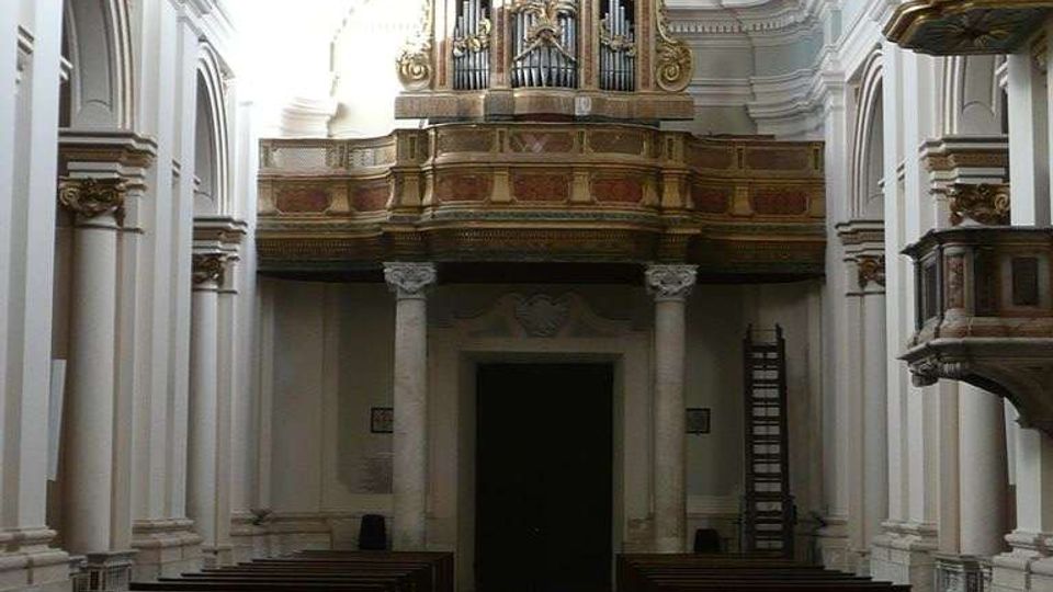 Interno di una chiesa con navata centrale e banchi in legno ai lati, pavimento a mosaico e alte arcate bianche. In fondo si vede una cantoria sopraelevata con un grande organo a canne decorato in oro.