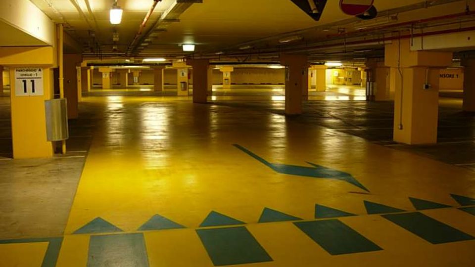 Parcheggio sotterraneo multipiano con pavimento giallo lucido e segnaletica direzionale blu, illuminato da lampade al neon. Pilastri numerati sostengono il soffitto basso con tubazioni a vista e cartelli stradali appesi.