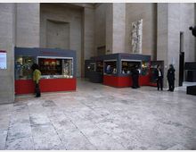 Ampio atrio interno di un edificio in pietra chiara con pavimento in lastre e pareti alte. Nello spazio sono presenti due chioschi con vetrine rosse e grigie per libreria e biglietteria, con una scultura murale in rilievo su una parete.