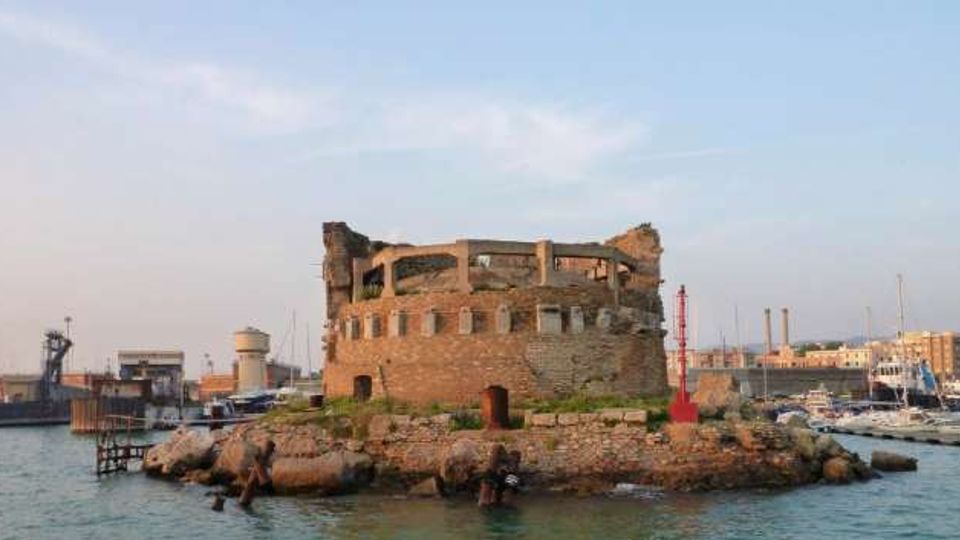 Ruderi di una fortificazione circolare in pietra su un piccolo isolotto roccioso all’ingresso di un porto. Intorno si vedono il mare, moli e strutture portuali con barche ormeggiate sullo sfondo.