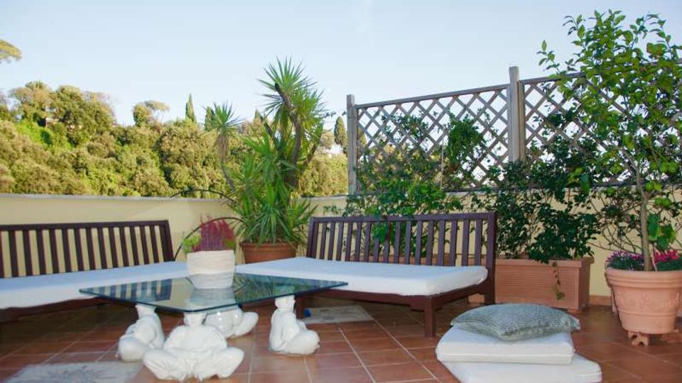 Terrazza esterna pavimentata in cotto con panche in legno e cuscini, tavolino basso con piano in vetro e base decorativa. Lungo il perimetro ci sono vasi con piante e una griglia a traliccio, con vista su alberi e cielo sullo sfondo.