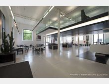 Ampio interno in stile industriale con pavimento chiaro, soffitto alto e soppalco con balaustra in vetro sostenuto da colonne. Lungo le pareti ci sono grandi finestre, illuminazione a binario e tavoli con sedie disposti come area ristorante o lounge.