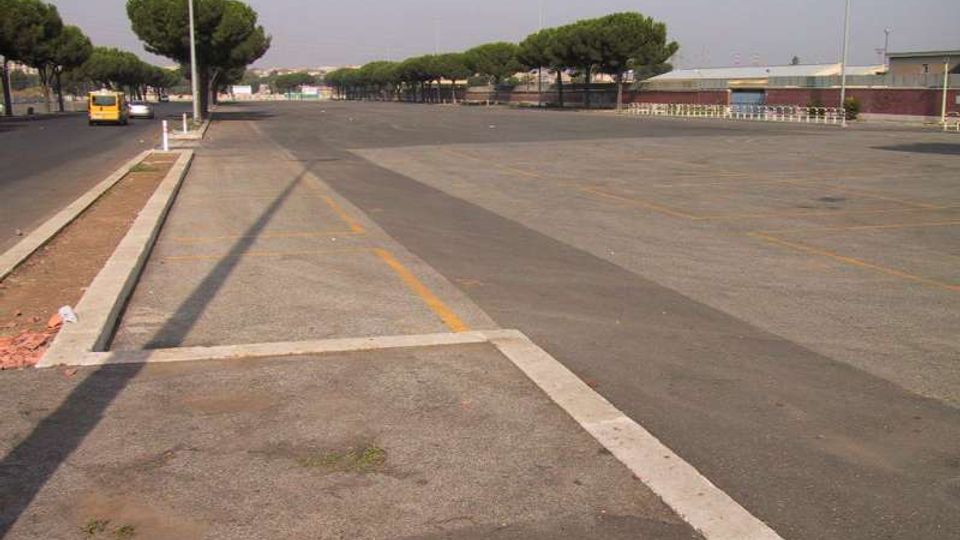 Ampio piazzale asfaltato con segnaletica orizzontale e corsie delimitate, affiancato da una strada con marciapiede e lampioni. Sullo sfondo si vedono filari di pini e alcuni edifici bassi in un’area periferica.