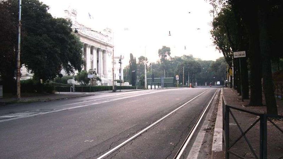 Ampio viale urbano con binari del tram al centro e marciapiede con ringhiera sul lato destro, fiancheggiato da alberi e lampioni. Sul lato sinistro si vede un grande edificio monumentale con facciata neoclassica a colonne e scalinata.