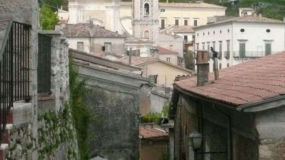 Vicolo in discesa tra edifici in pietra e intonaco con tetti in coppi, che conduce verso il centro di un borgo. Sullo sfondo si vedono una chiesa con facciata classica e un campanile, con colline boscose tutt’intorno.