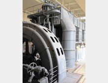Grande sala industriale con un enorme macchinario metallico grigio, con carter curvo forato, tubazioni e passerella superiore con ringhiera. Sullo sfondo si vedono pannelli e condotti di grandi dimensioni allineati lungo la parete, con pavimento in piastrelle e griglie di aerazione.