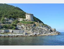 Torre costiera in pietra di forma cilindrica con merlature, situata su una scogliera rocciosa ai piedi di una collina verde. In primo piano si vede il mare con una scogliera e un basso muro di contenimento lungo la riva.